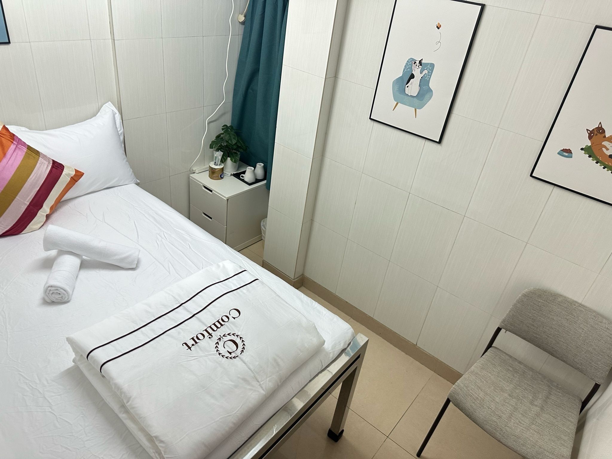 Ein ordentlich gemachtes Bett mit weißer Decke und einem zusammengerollten Handtuch, ein Nachttisch mit Pflanze, ein gerahmtes Bild an der Wand und ein Sessel in der Ecke.