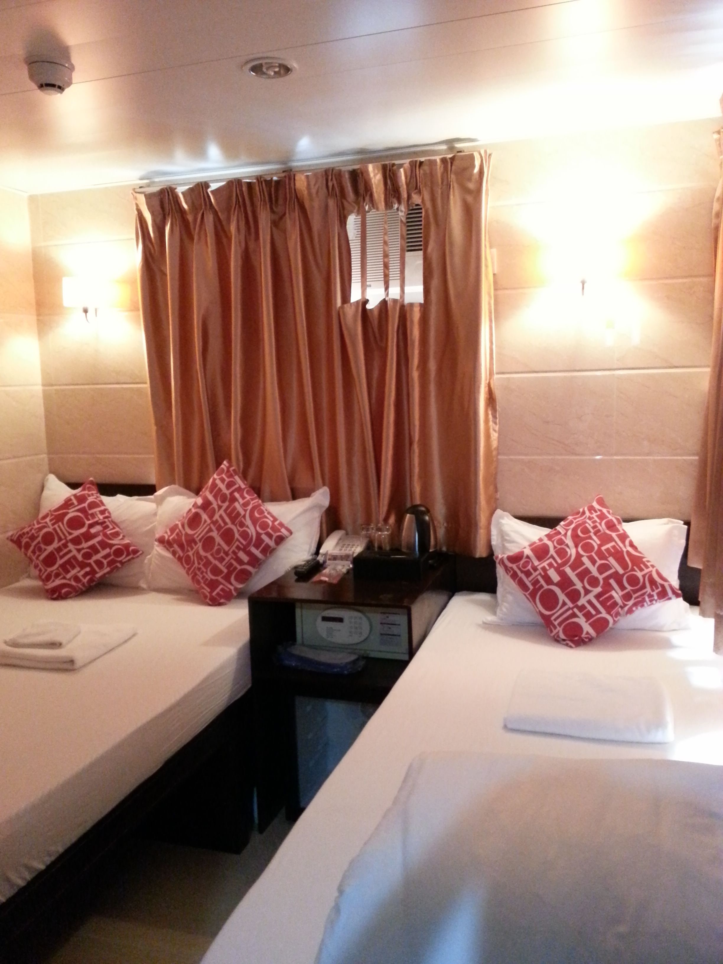 Une chambre d’hôtel avec deux lits, chacun agrémenté de draps blancs et d’oreillers à motifs rouges. Une petite table avec un téléphone et une machine à café se trouve entre les lits. Les murs de la chambre sont de couleur beige et il y a une fenêtre avec des rideaux.