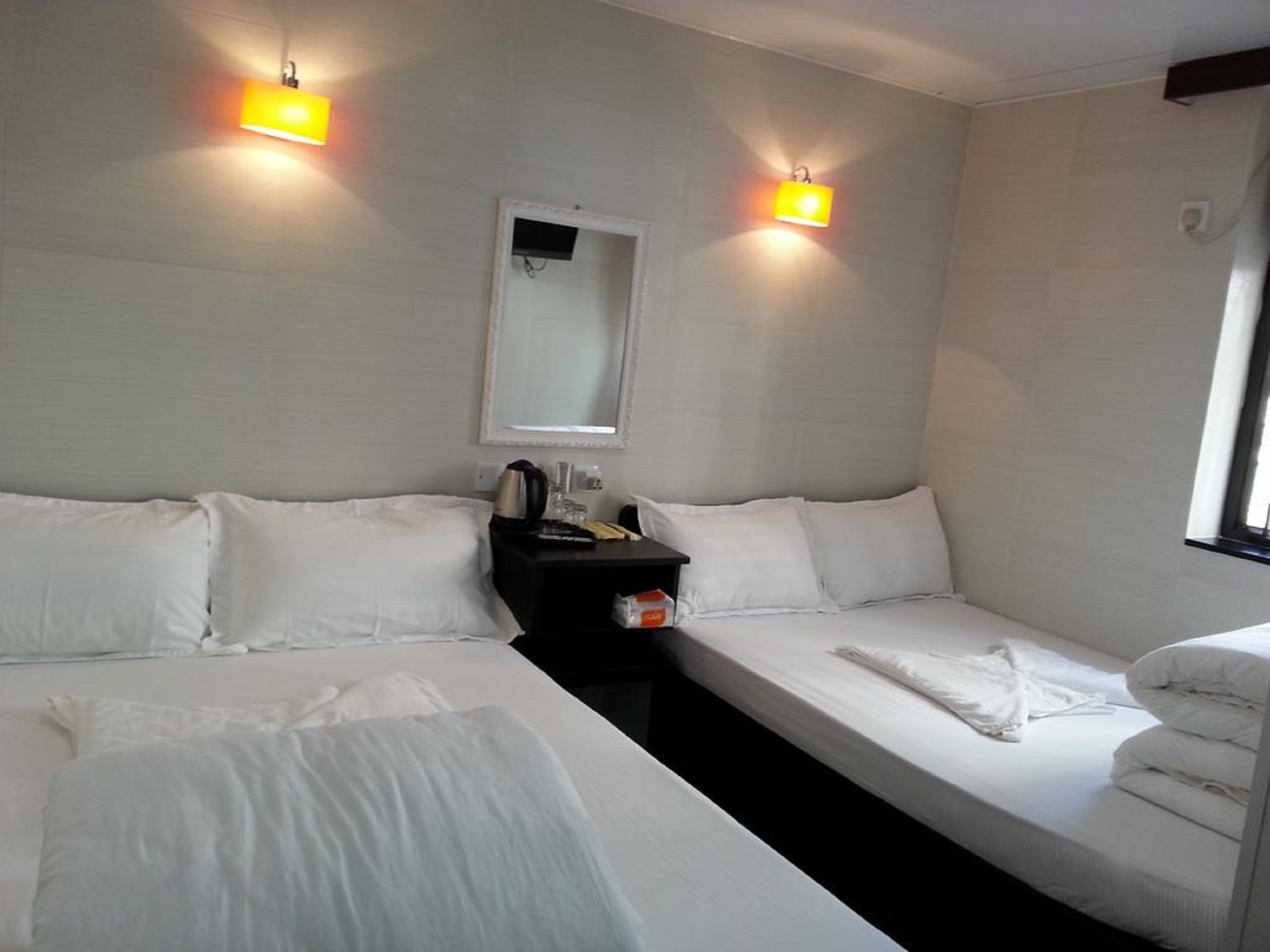 A hotel room with two single beds, a small bedside table with a kettle, a mirror, and two wall-mounted lights.