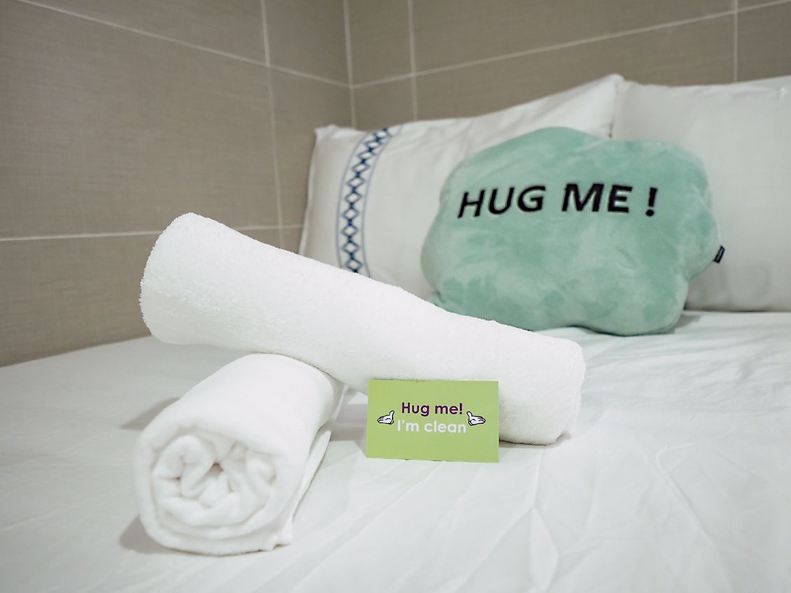 A neatly made bed with a rolled white towel and a pillow with the text "HUG ME!".
