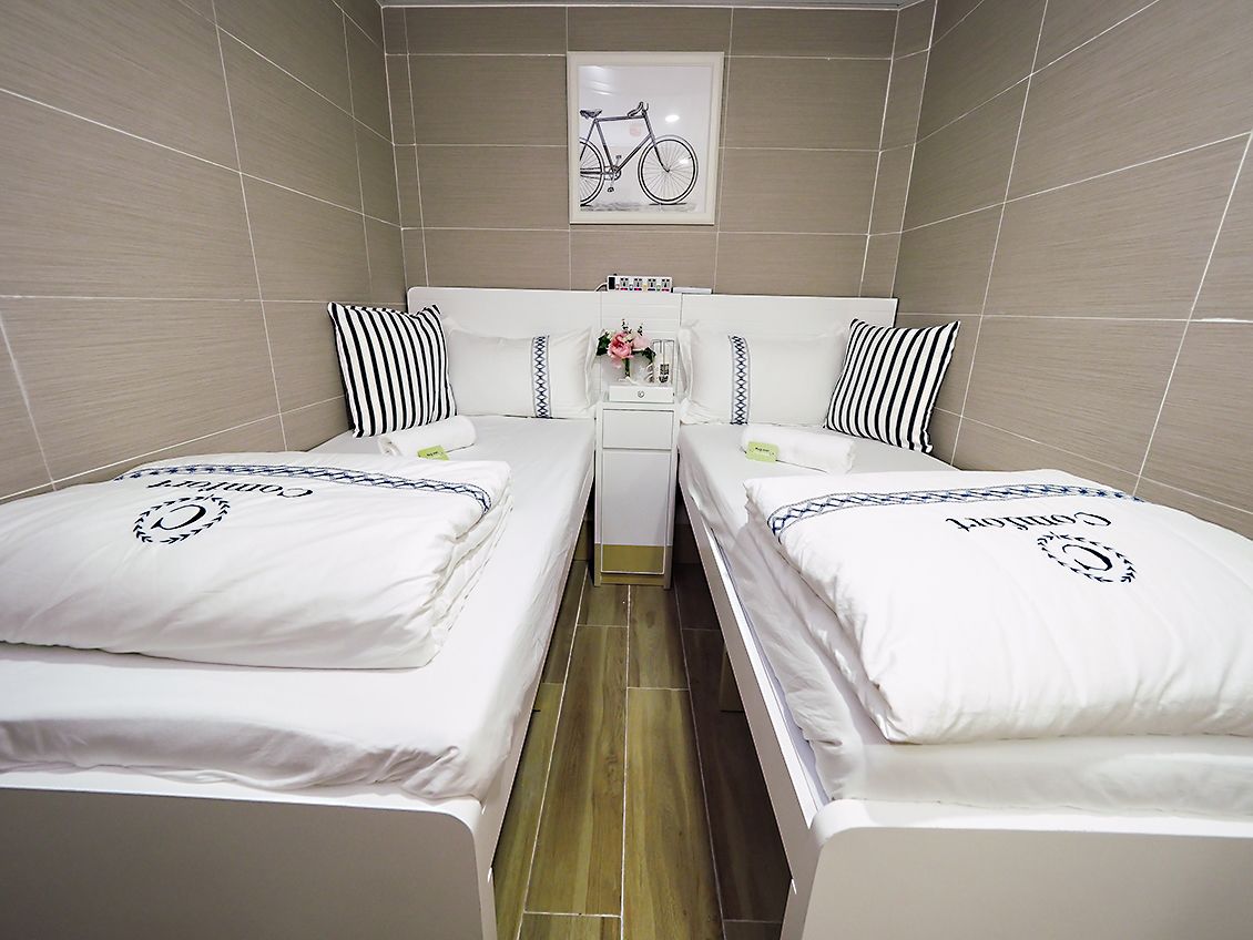 A room with two beds, each with a pillow featuring a monogram. There is a small table with a vase of flowers between the beds.