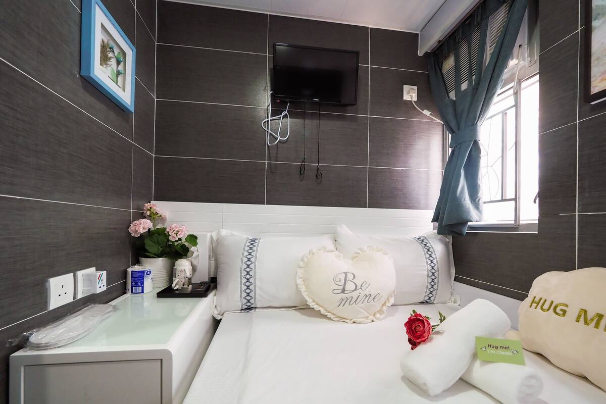 A hotel room with a bed, a bedside table, a TV mounted on the wall, and a window with curtains.