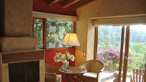 Une pièce chaleureuse avec une cheminée, un fauteuil en osier, une petite table avec une lampe et une vue sur une végétation luxuriante, visible à travers de grandes fenêtres.