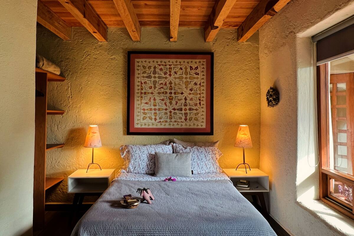 Ein Schlafzimmer mit Holzbalkendecke, einem großen Wandbehang mit Muster, zwei Nachttischlampen, einem Bett mit floralem Überwurf und einem Fenster mit Blick.