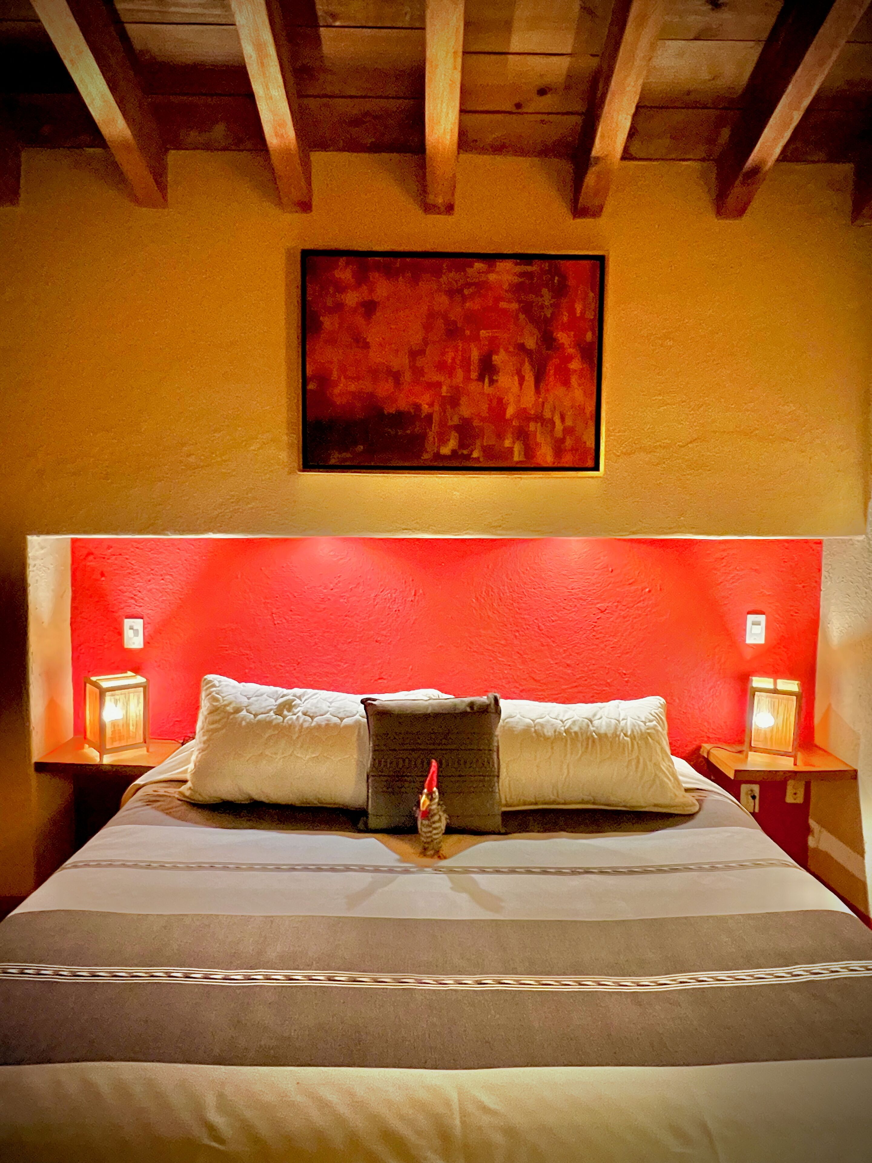 A bed with white linens and two pillows, a red accent wall, and a framed abstract painting above the bed.