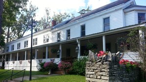 A white two-story building with a covered porch, stone wall, and hanging flower baskets. - The Baseball Hotel (Richfield Springs)