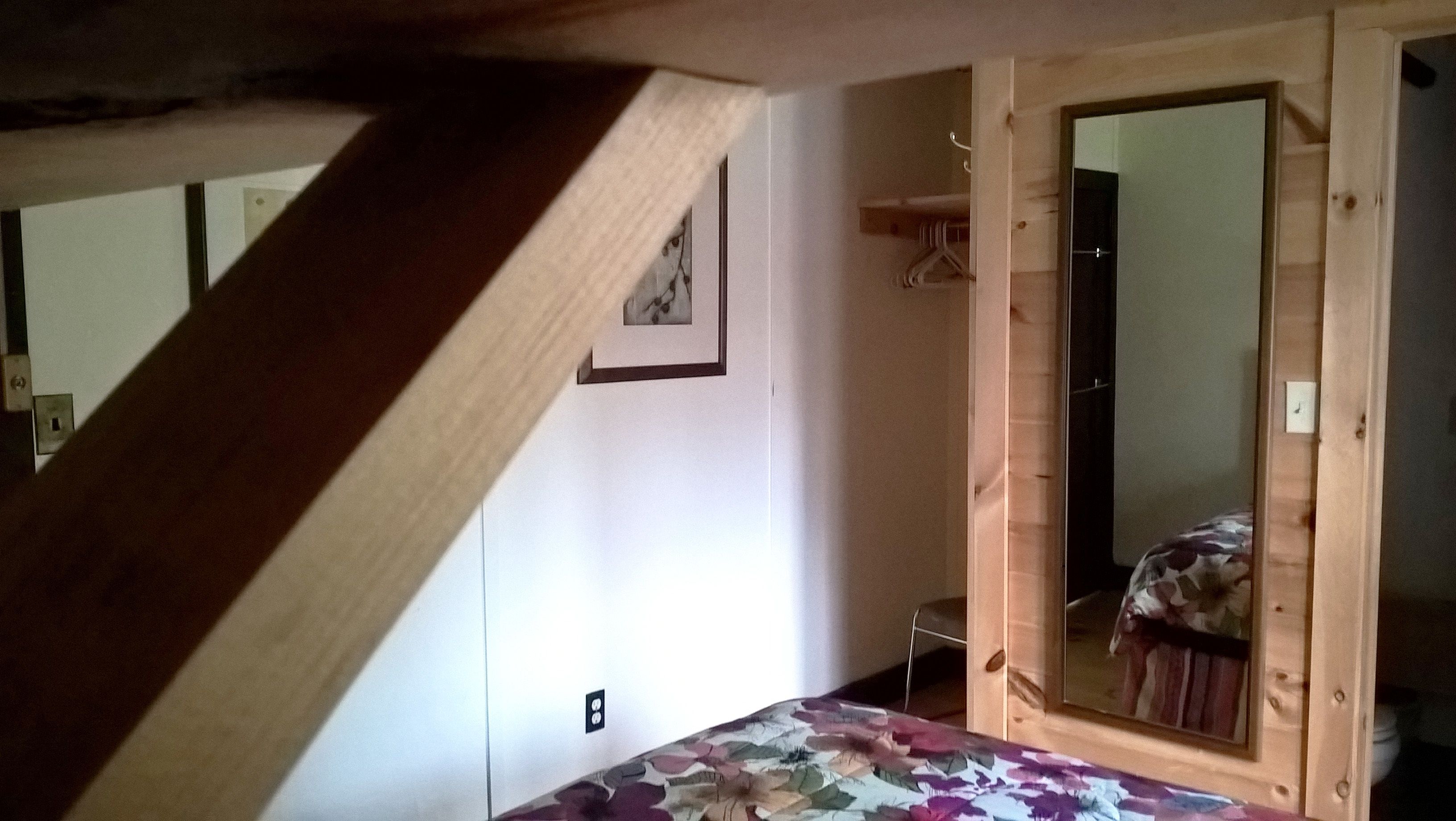 Een slaapkamer met een houten trap, een bed met een bloemenprint deken, een kledingkast en een spiegel die een deel van de kamer weerspiegelt.