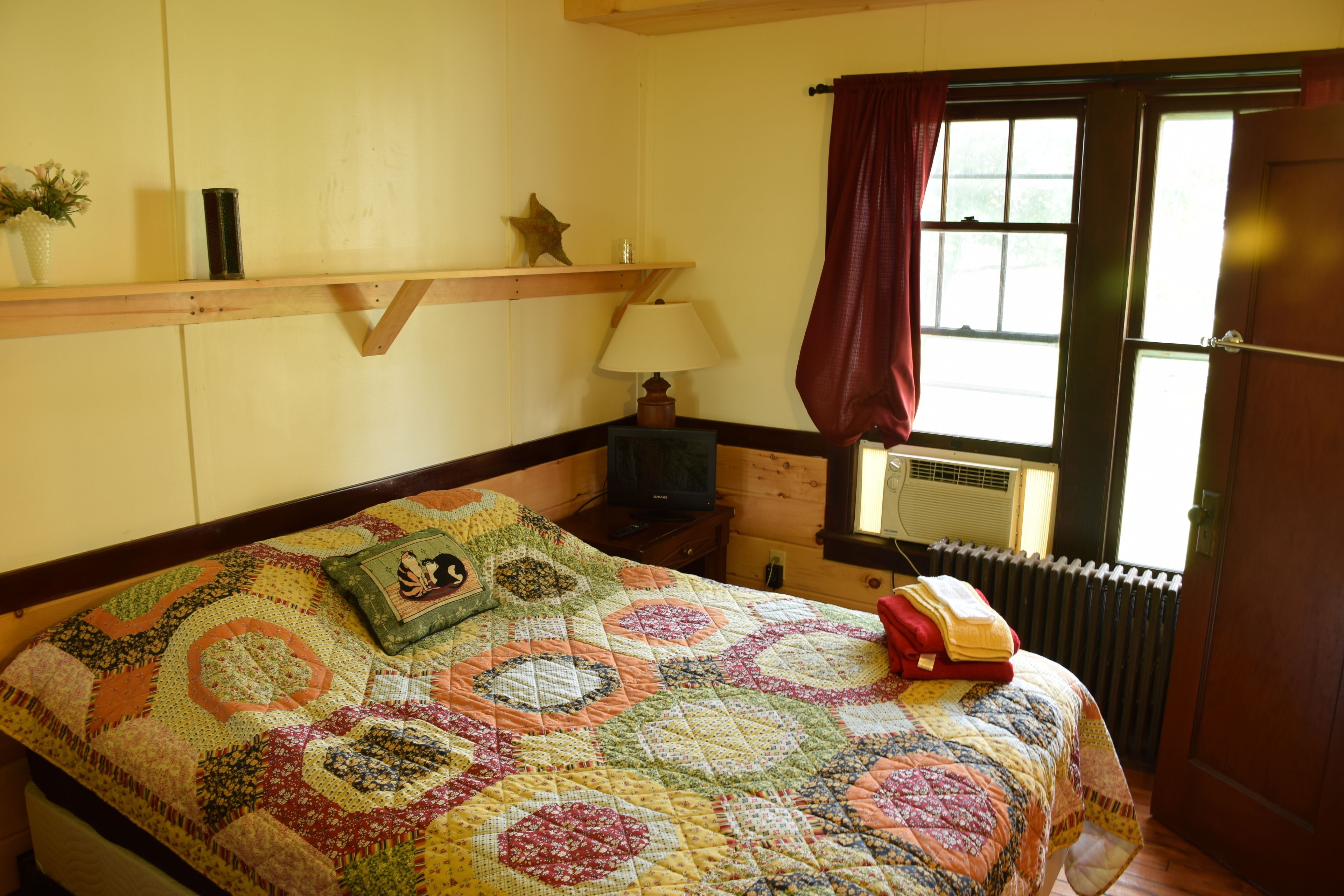 A bedroom with a large bed, a wooden shelf with decorative items, a window with curtains, and a radiator.