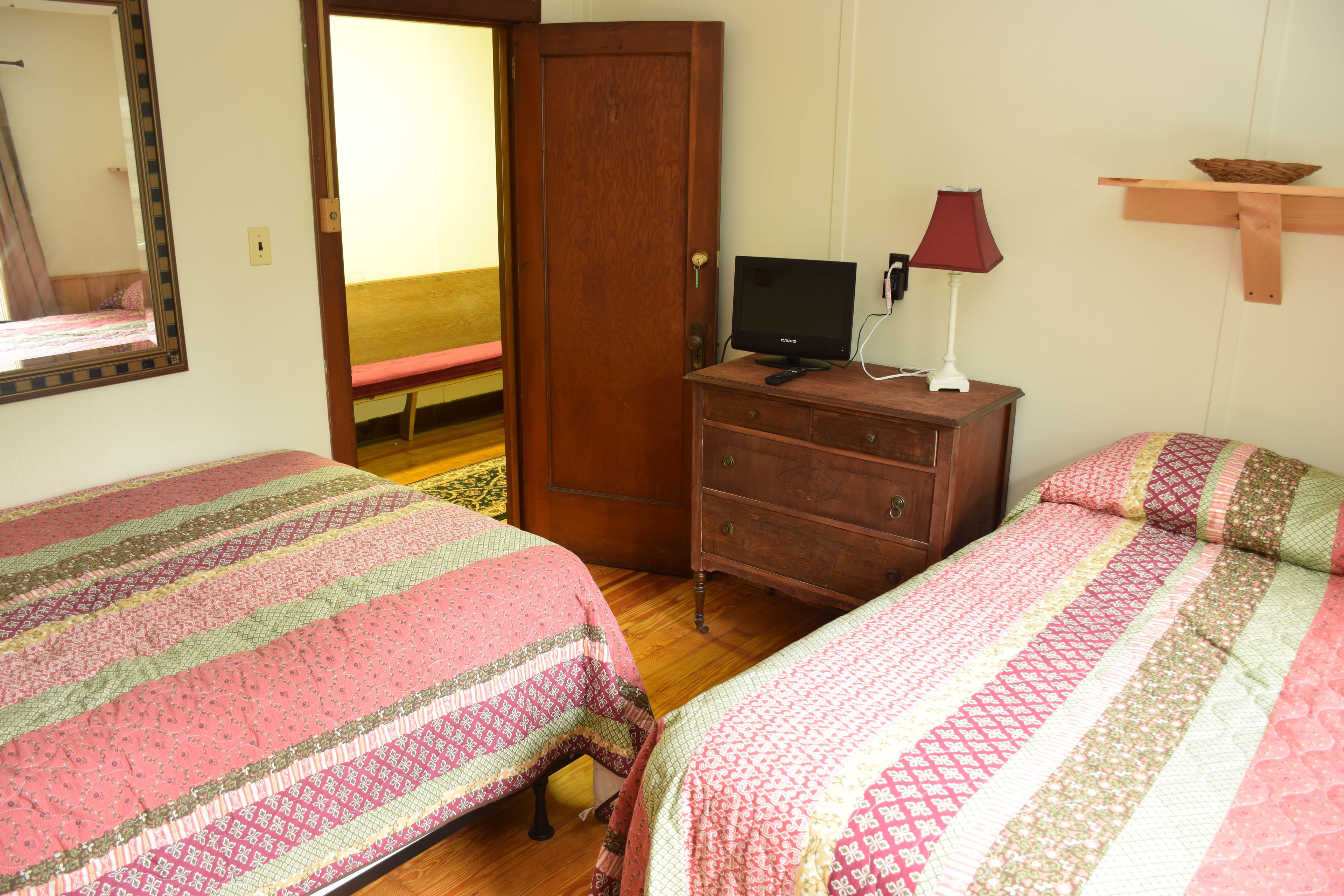 Quarto com duas camas, um aparador de madeira, uma televisão sobre o aparador e um espelho na parede.