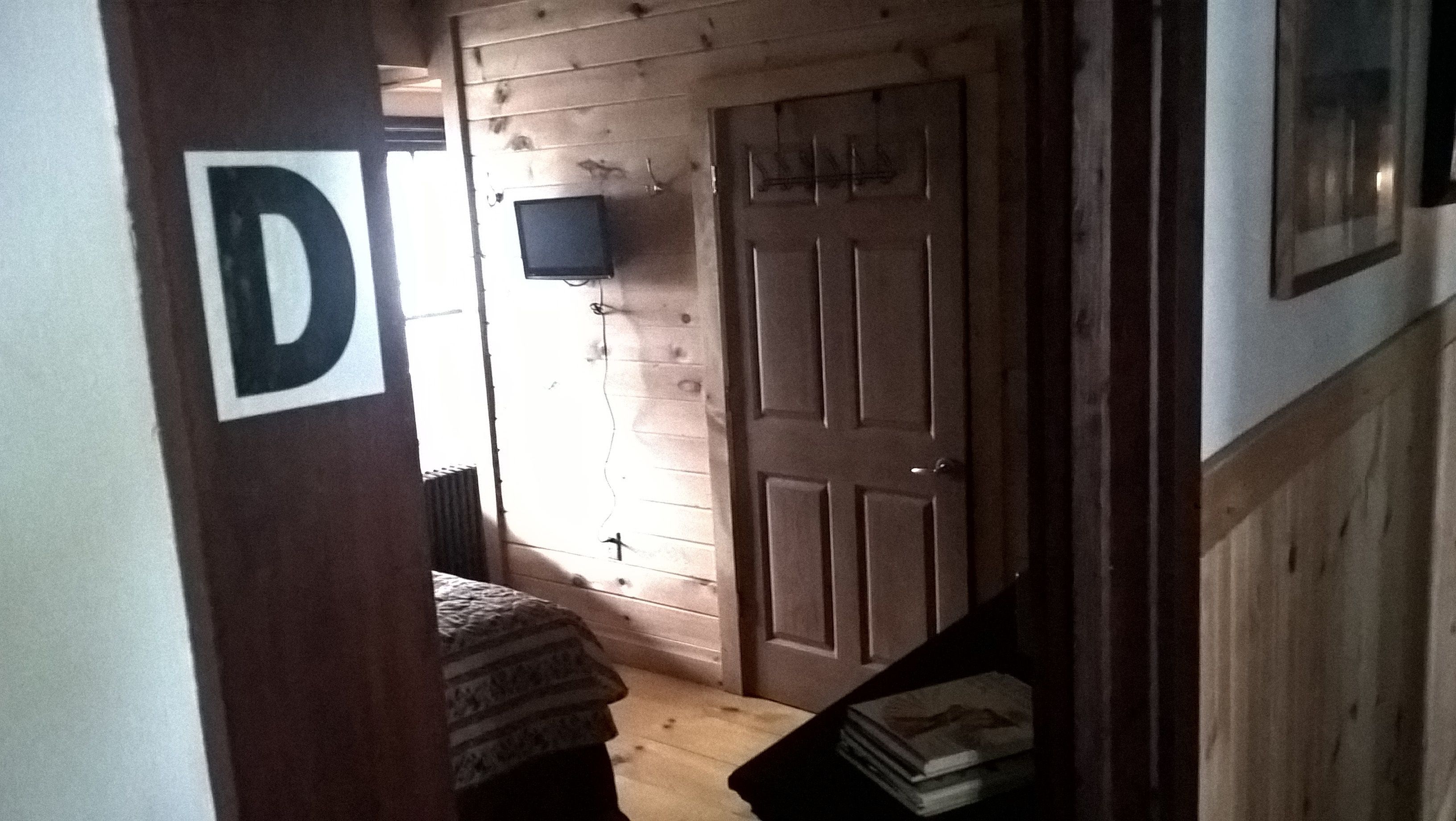 A narrow hallway with a wooden door, a television mounted on the wall, and a bed with a patterned blanket.