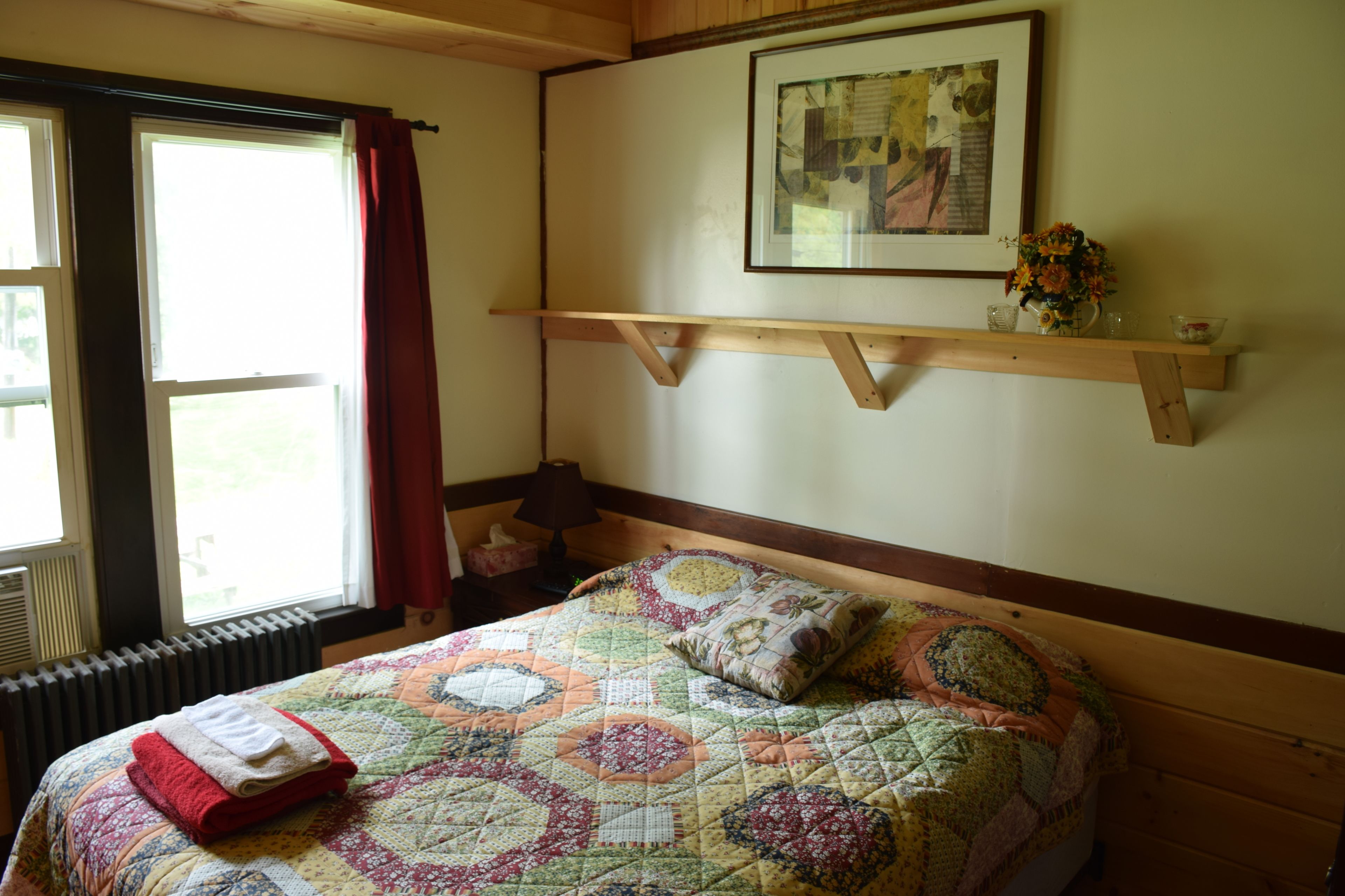 Een slaapkamer met een houten bed, een sprei, een rode handdoek, een raam met gordijnen, een wandplank met een vaas en een lijst met een afbeelding.