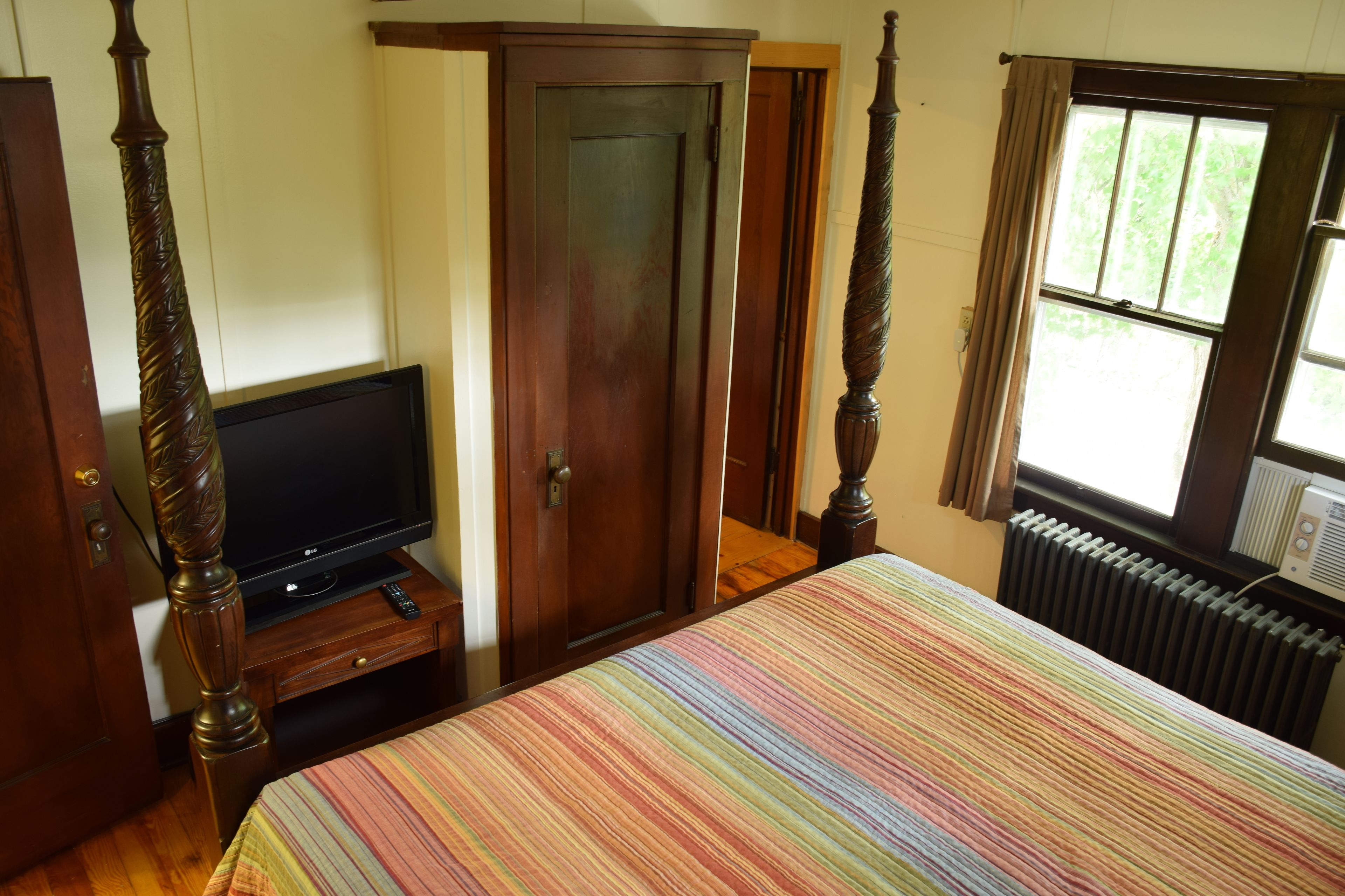 A bedroom with a bed, a television on a wooden stand, a wardrobe, and a window with curtains.