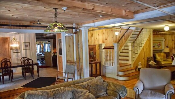A cozy living room with a wooden ceiling, a sofa, a chair, and a staircase leading to another level. - The Baseball Hotel (Richfield Springs)