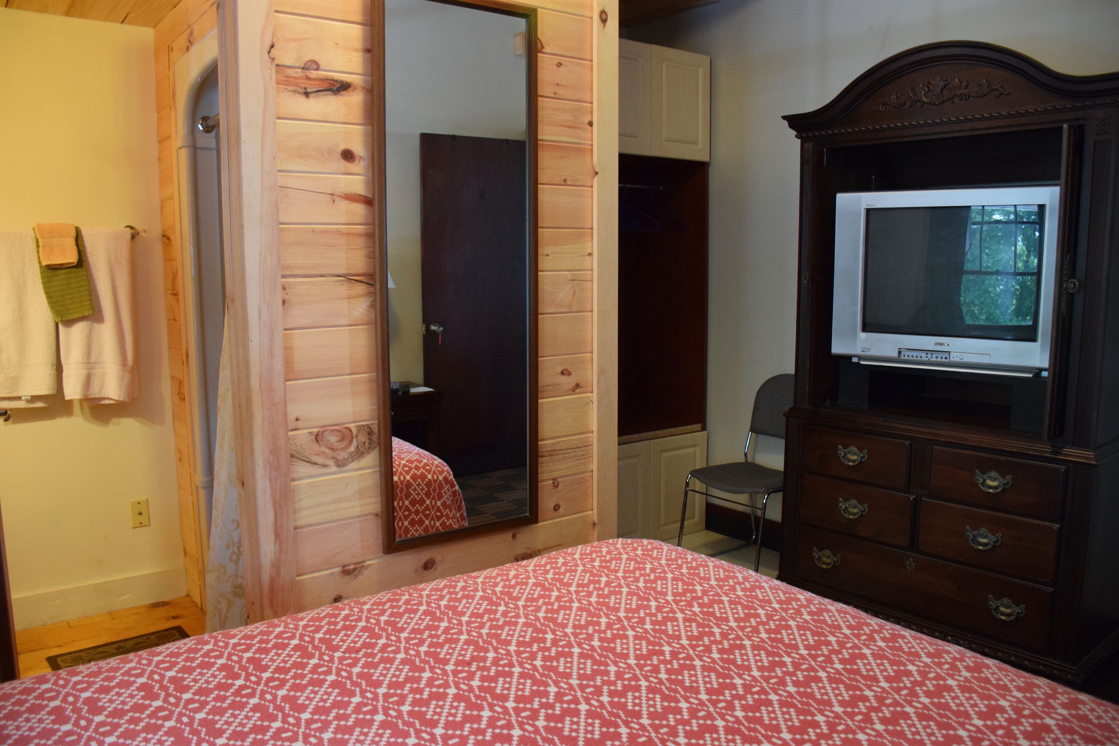 A bedroom with a wooden wardrobe, a bed with a patterned bedspread, a mirror, and a television on a dresser.