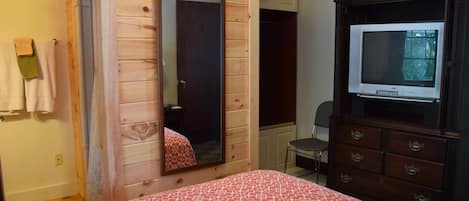 A bedroom with a wooden wardrobe, a bed with a patterned bedspread, a mirror, and a television on a dresser.