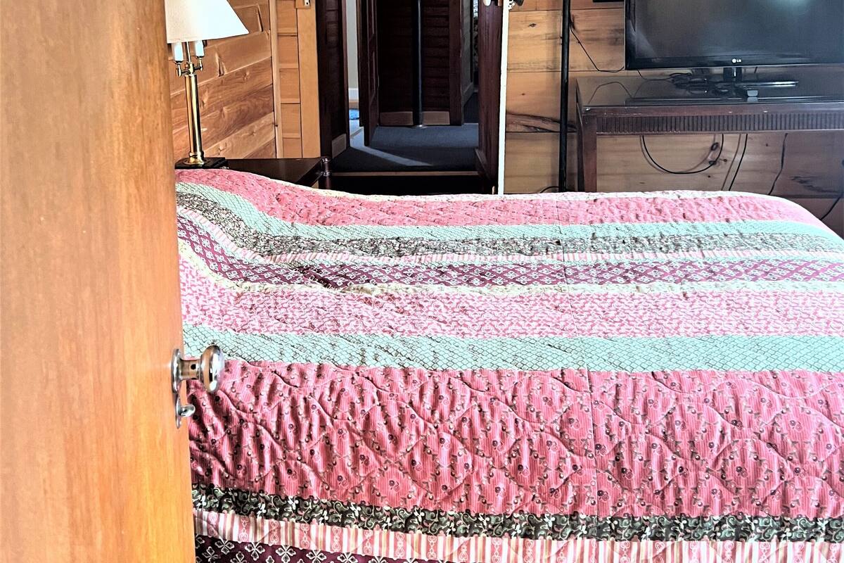 A cozy attic bedroom with a wooden ceiling, a bed with a patterned quilt, a television, and a door leading to another room.