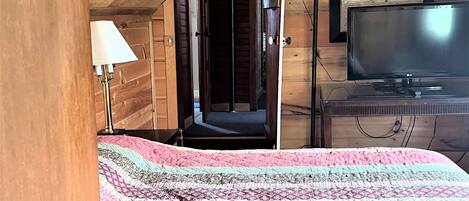 A cozy attic bedroom with a wooden ceiling, a bed with a patterned quilt, a television, and a door leading to another room.
