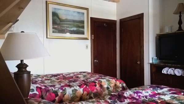 A bedroom with a floral bedspread, a painting on the wall, a lamp, and a TV on a wooden cabinet. - The Baseball Hotel (Richfield Springs)