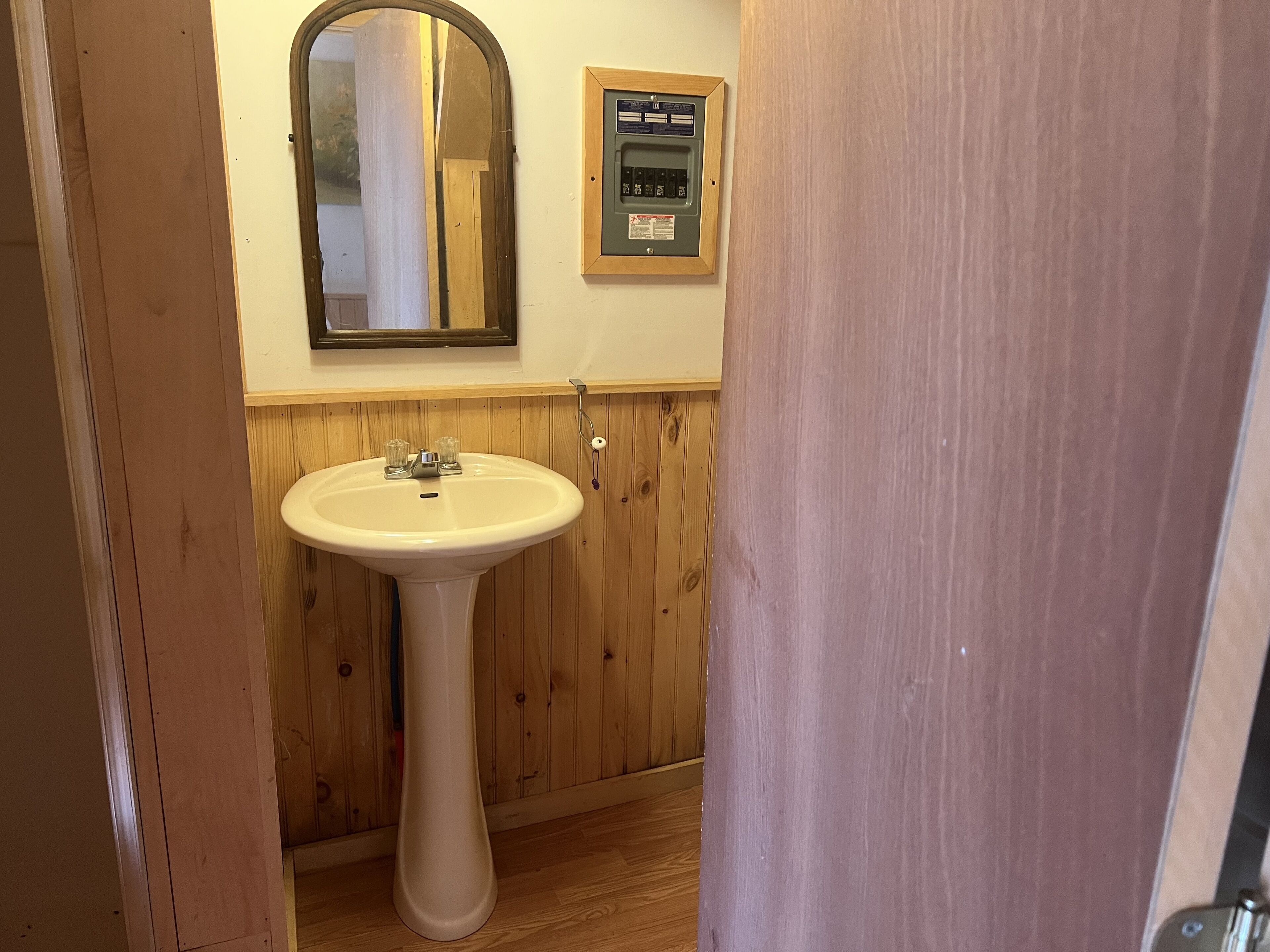 A bathroom with a pedestal sink, wooden paneling, a mirror, and a wall-mounted electrical panel.