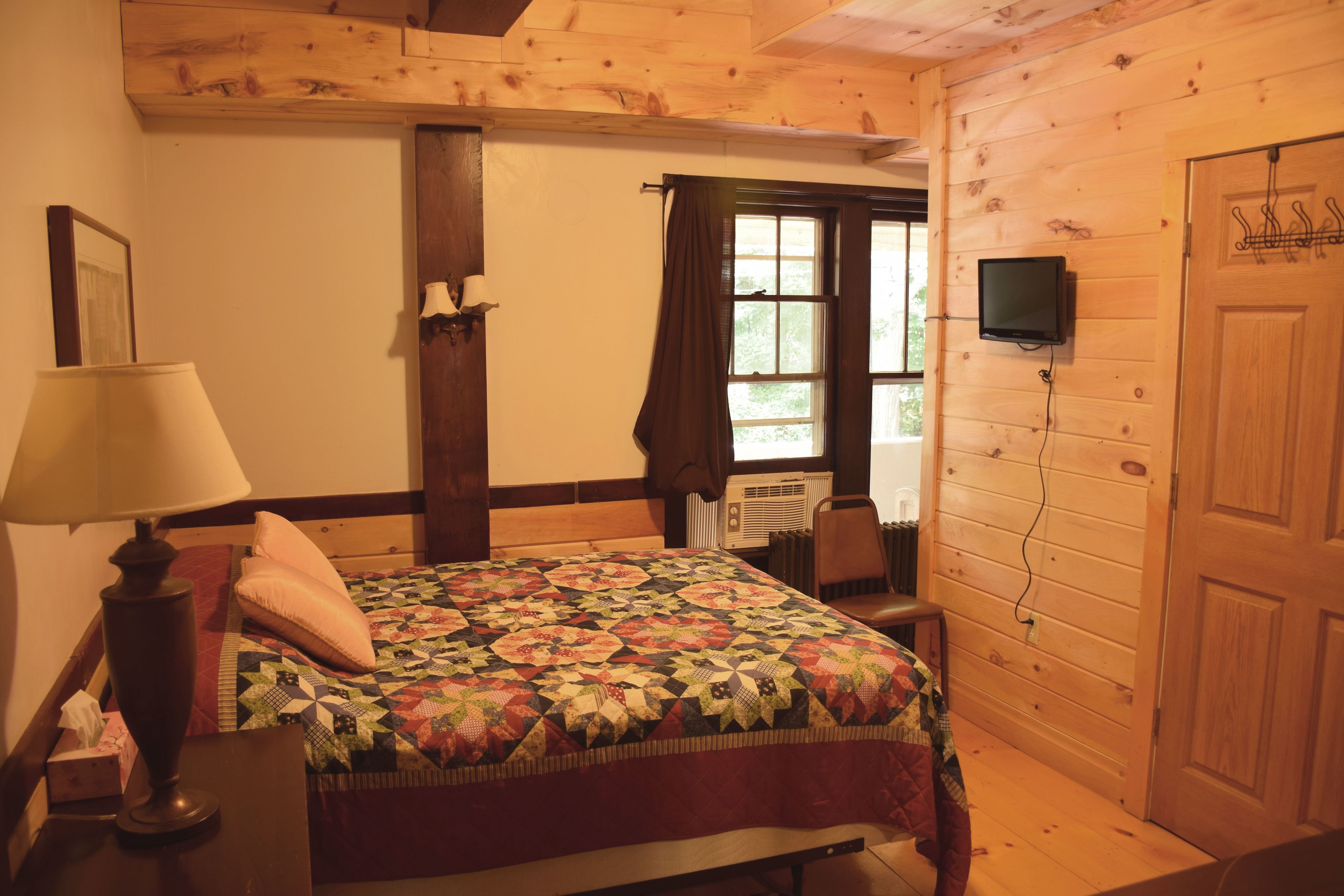 A cozy room with a wooden ceiling, a bed with a colorful quilt, a bedside lamp, a TV on the wall, and a door with a coat rack.