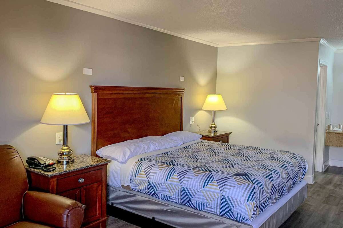 A hotel room with a bed, two bedside lamps, a telephone, a leather chair, and a bathroom with a sink and mirror.