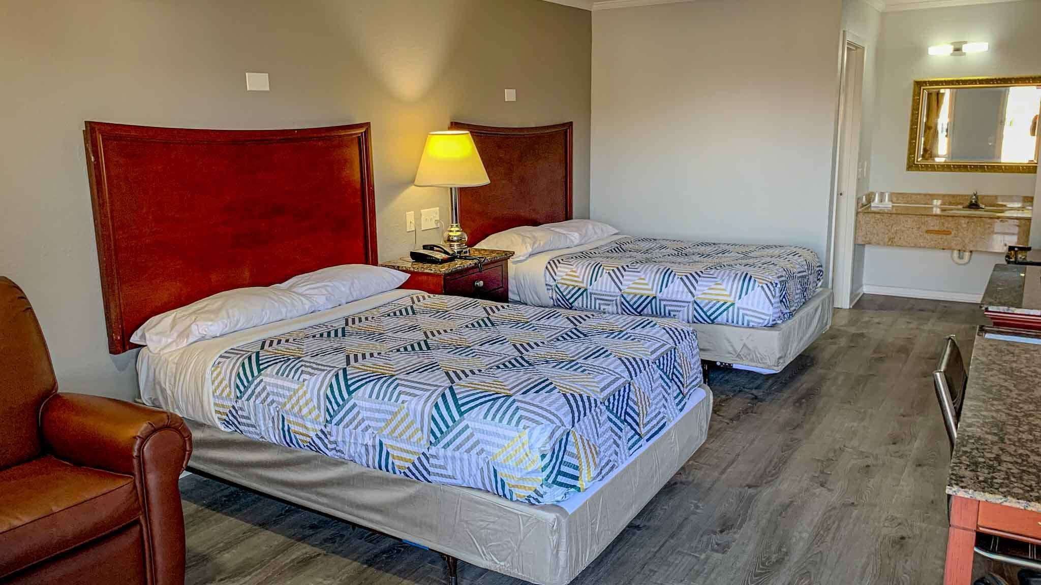 Habitación de hotel con dos camas, cabecero de madera, mesita de noche con lámpara, un escritorio pequeño y un baño con espejo y lavamanos.