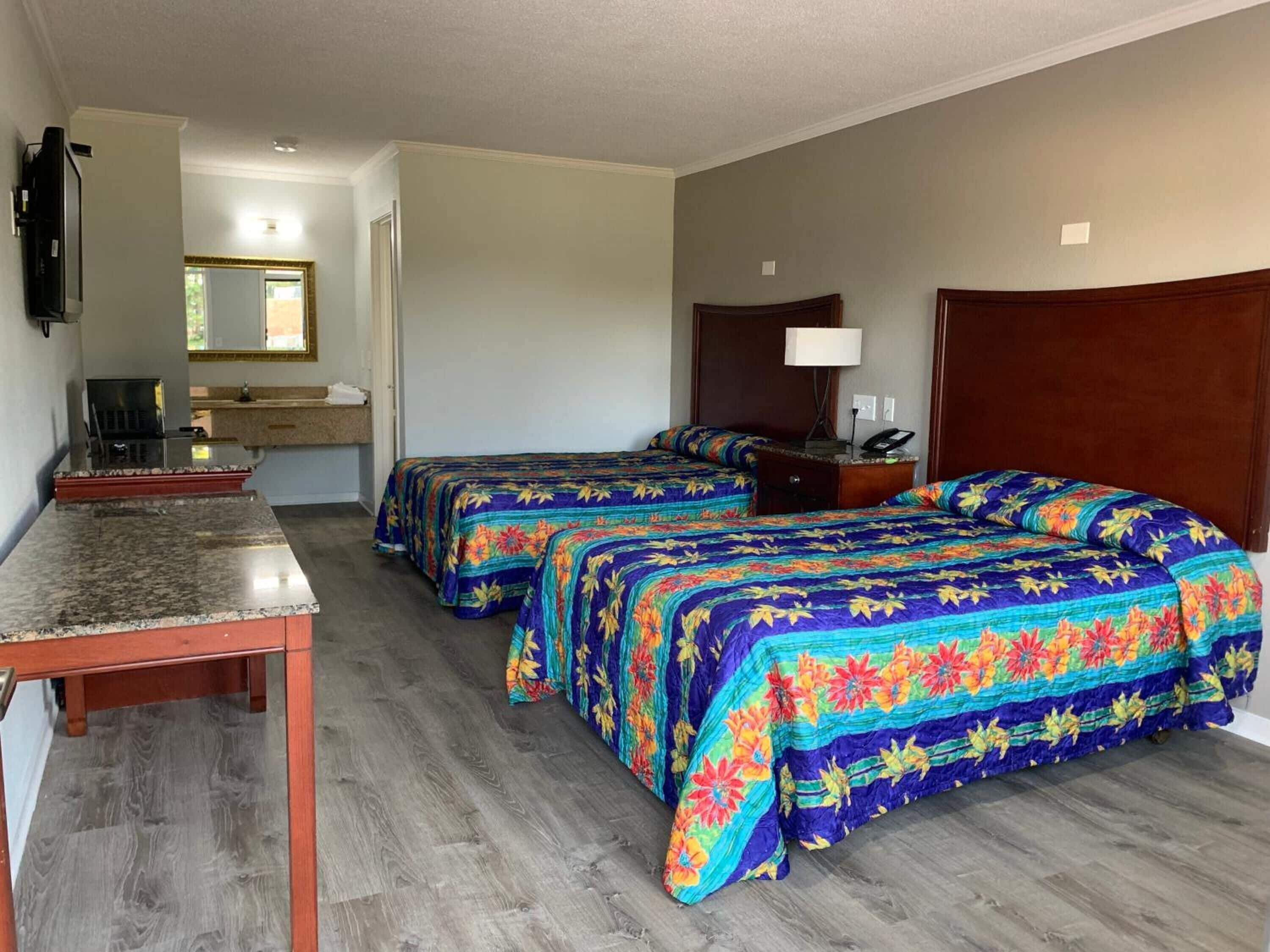 A hotel room with two beds, a desk, a television, a mirror, and a bathroom visible in the background.