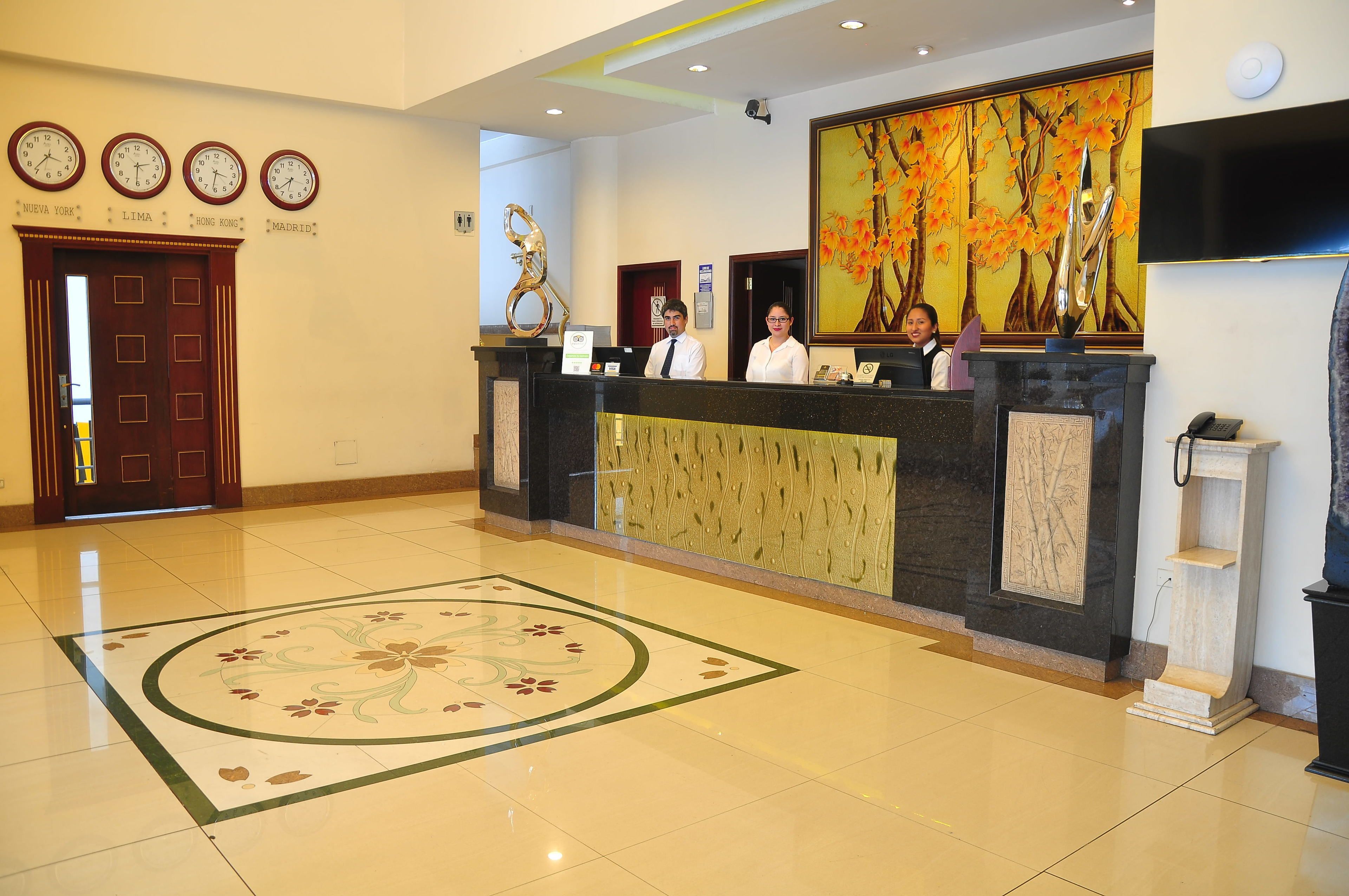 A hotel reception area with a reception desk, clocks displaying times for New York, Lima, Hong Kong, and Madrid, and a large painting of autumn leaves.