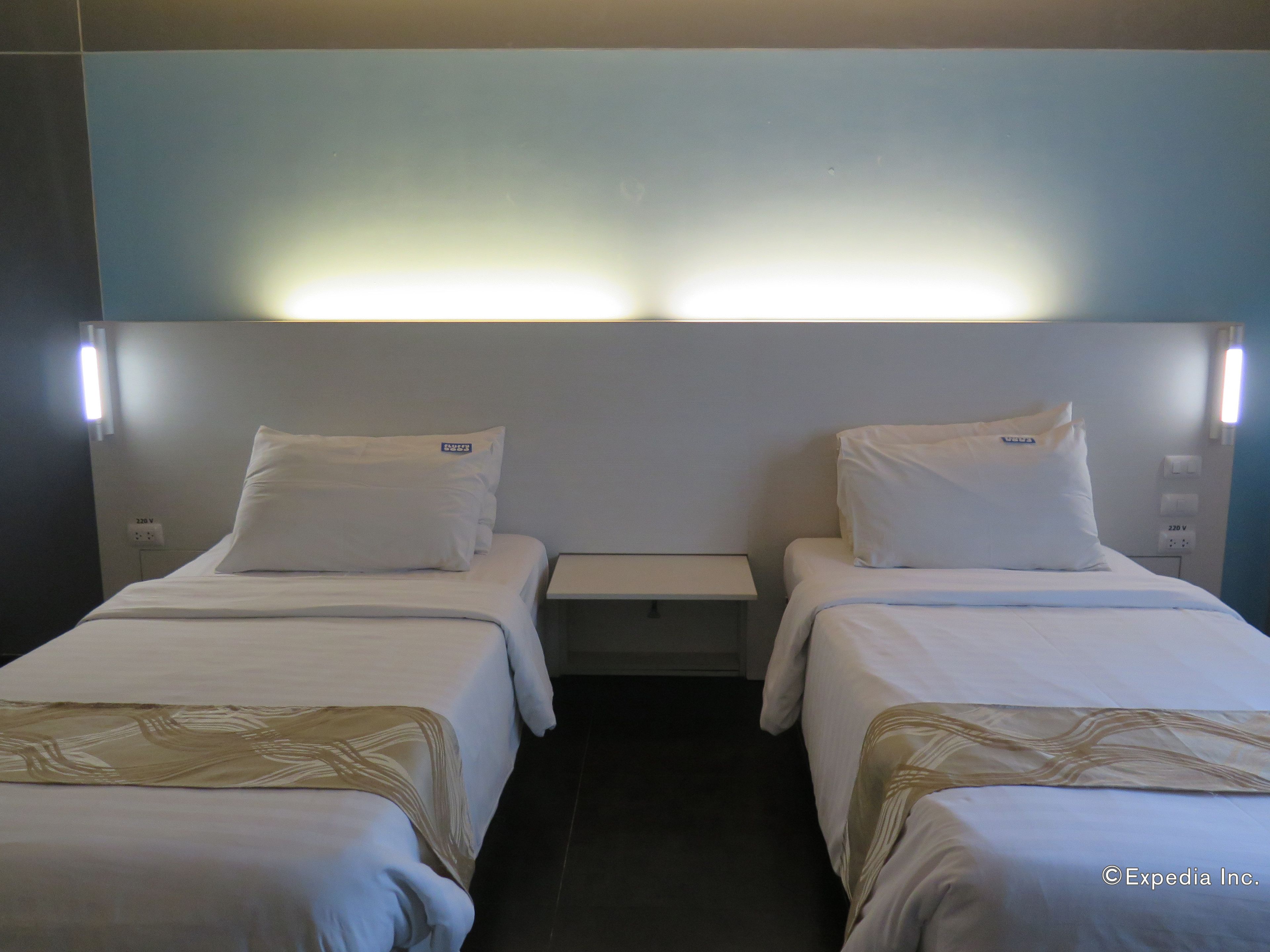 Habitación de hotel con dos camas individuales, cada una con un almohadón blanco y cabecero. Hay una mesita de noche pequeña entre las camas, y una lámpara de pared sobre ellas.