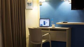 Quarto de hotel com uma cama, uma escrivaninha com um laptop, uma cadeira e uma TV fixada na parede.