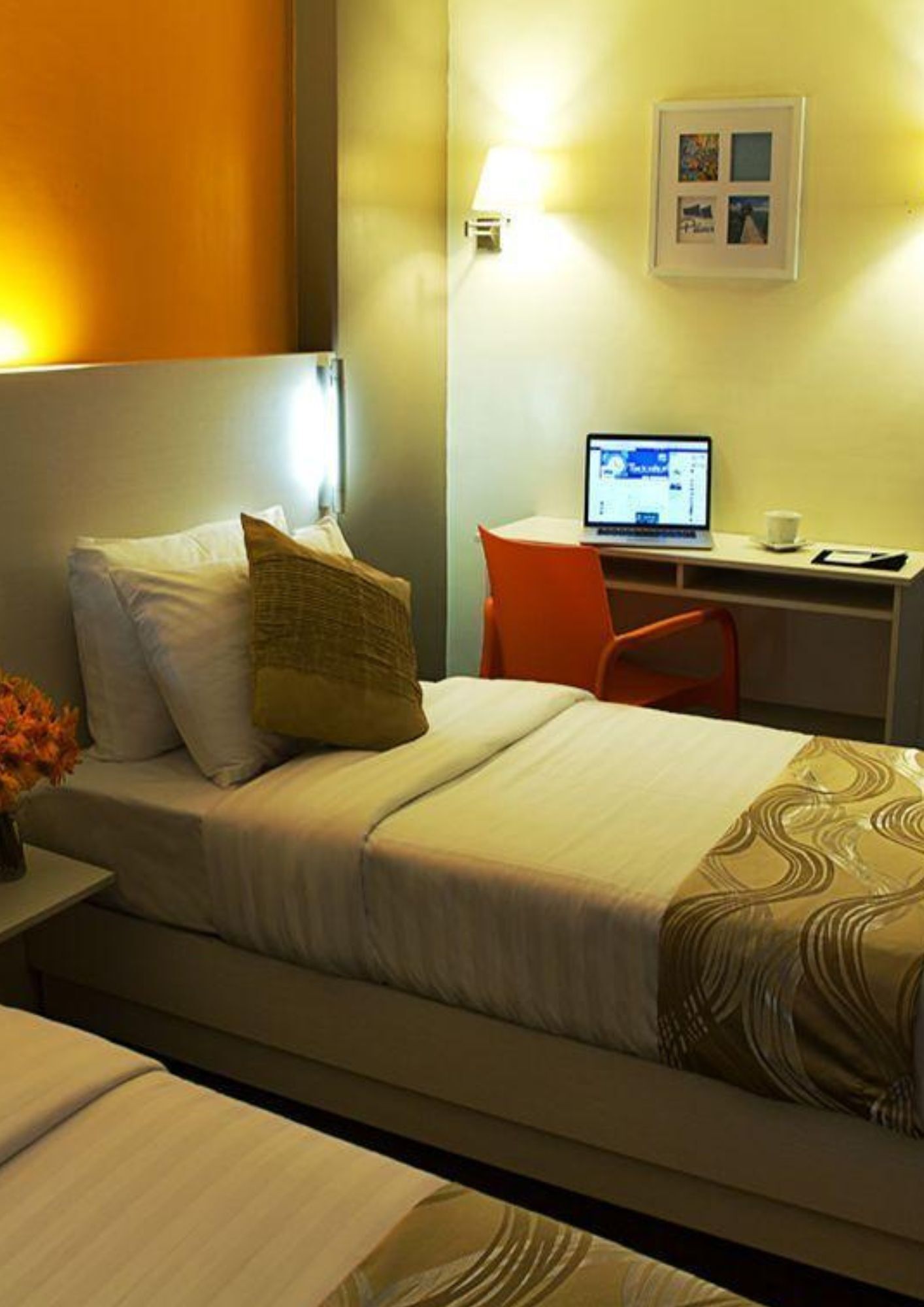 A hotel room with a bed, a desk with a laptop, a chair, and a small table with a vase of flowers.