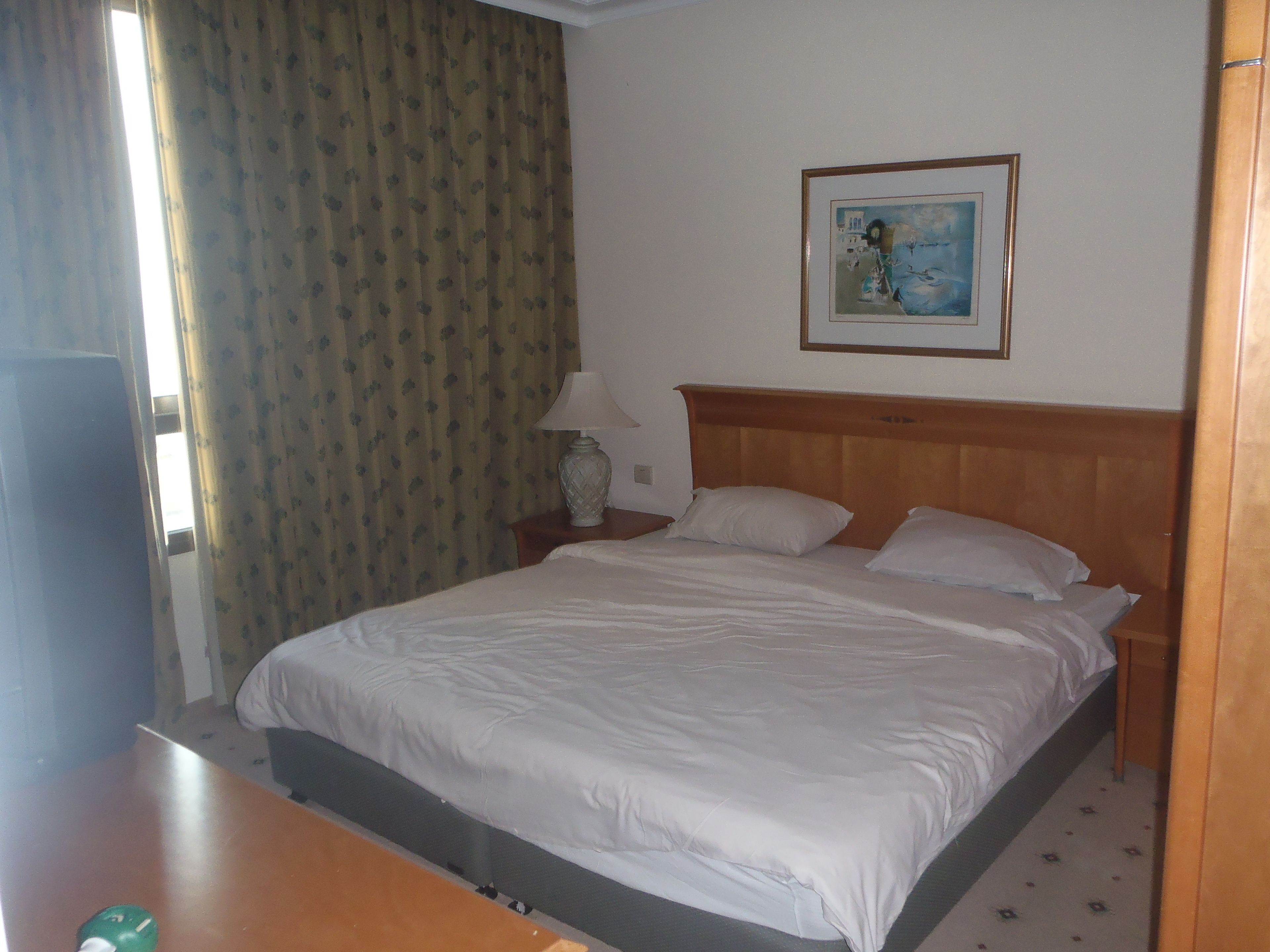 Una habitación de hotel con una cama, una mesita de noche, una lámpara, un cuadro en la pared y una ventana con cortinas.