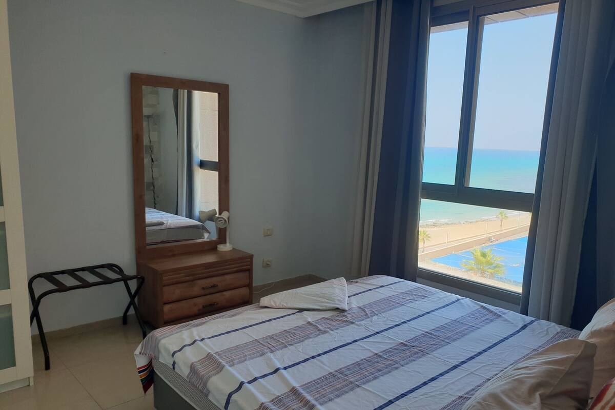 Une chambre à coucher avec un lit, une commode, un miroir et une fenêtre donnant sur la plage.