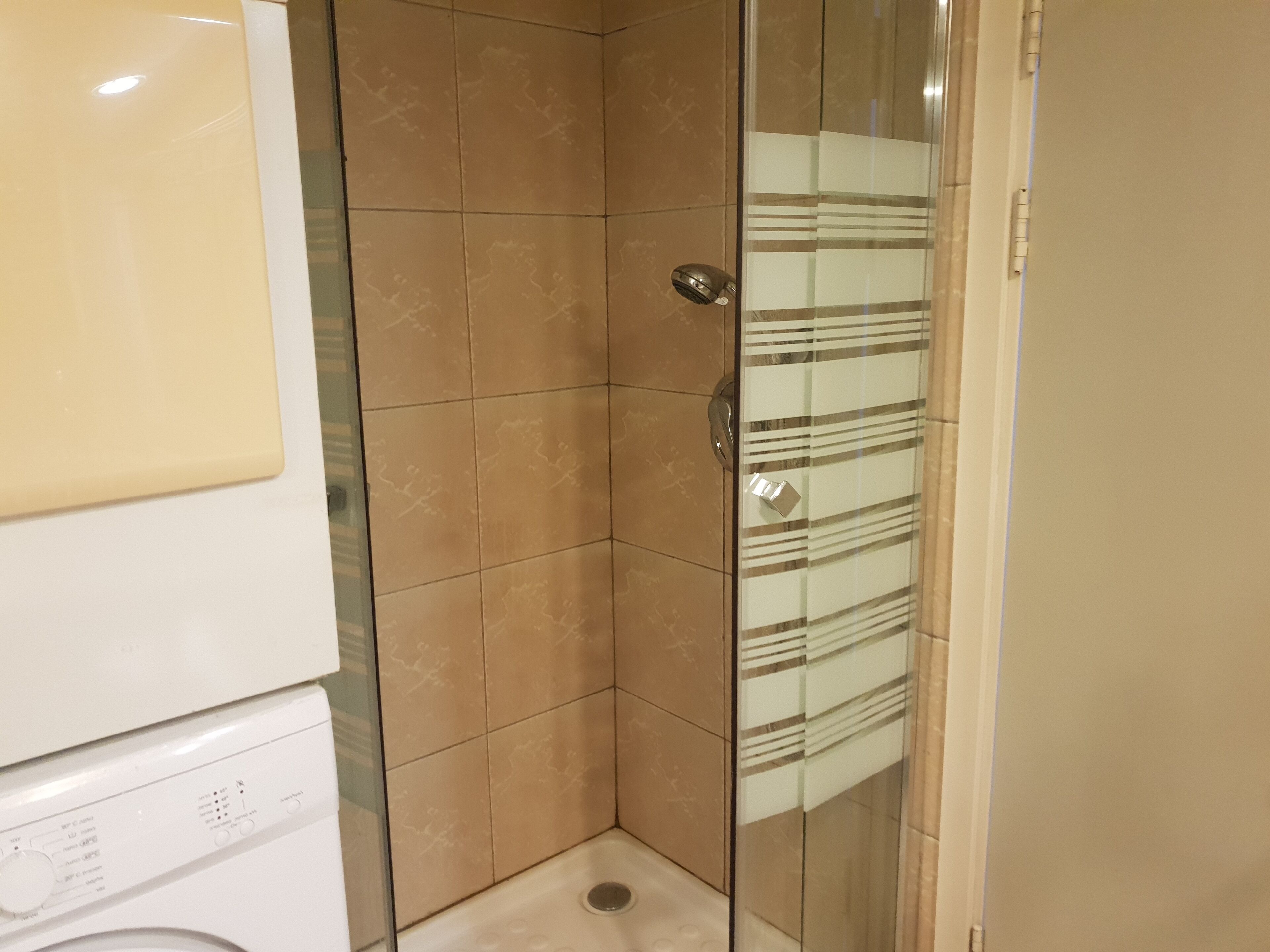A bathroom with a glass shower door, a white washing machine, and beige tiles.