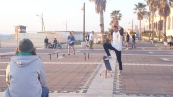 En offentlig plads med en skateboarder der udfører et trick, folk der sidder på bænke, og palmer i baggrunden.