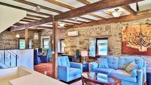 A spacious living area with a stone wall, wooden beams, and blue upholstered furniture. - All Suites Whitney Manor (Kingston)