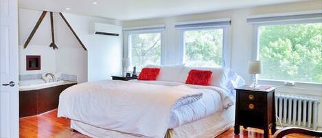 A spacious bedroom with a large bed, a wooden floor, a cowhide rug, and a view of trees through the windows.