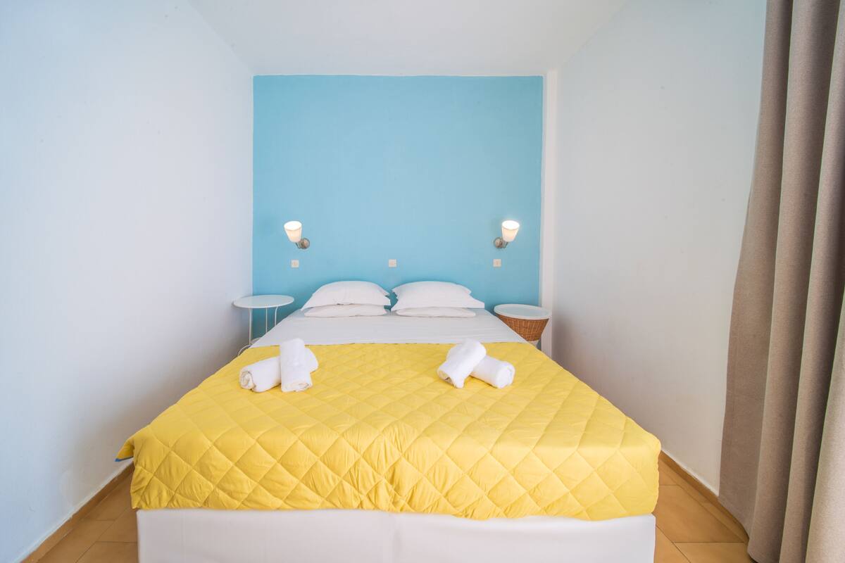 Una cama bien hecha con colcha amarilla, almohadas blancas y dos toallas enrolladas sobre un cabecero blanco.