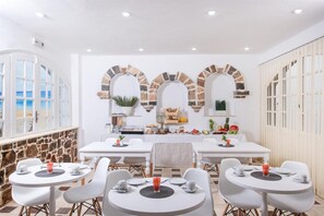 Un comedor con mesas y sillas blancas, una pared de piedra y vista a la playa.