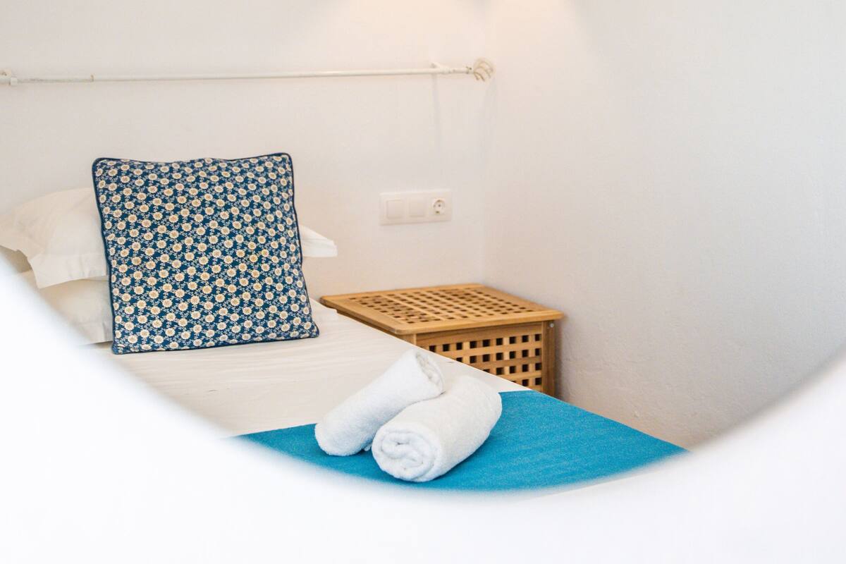 Un dormitorio con cama, almohada azul, mesita de madera y lámpara colgante.