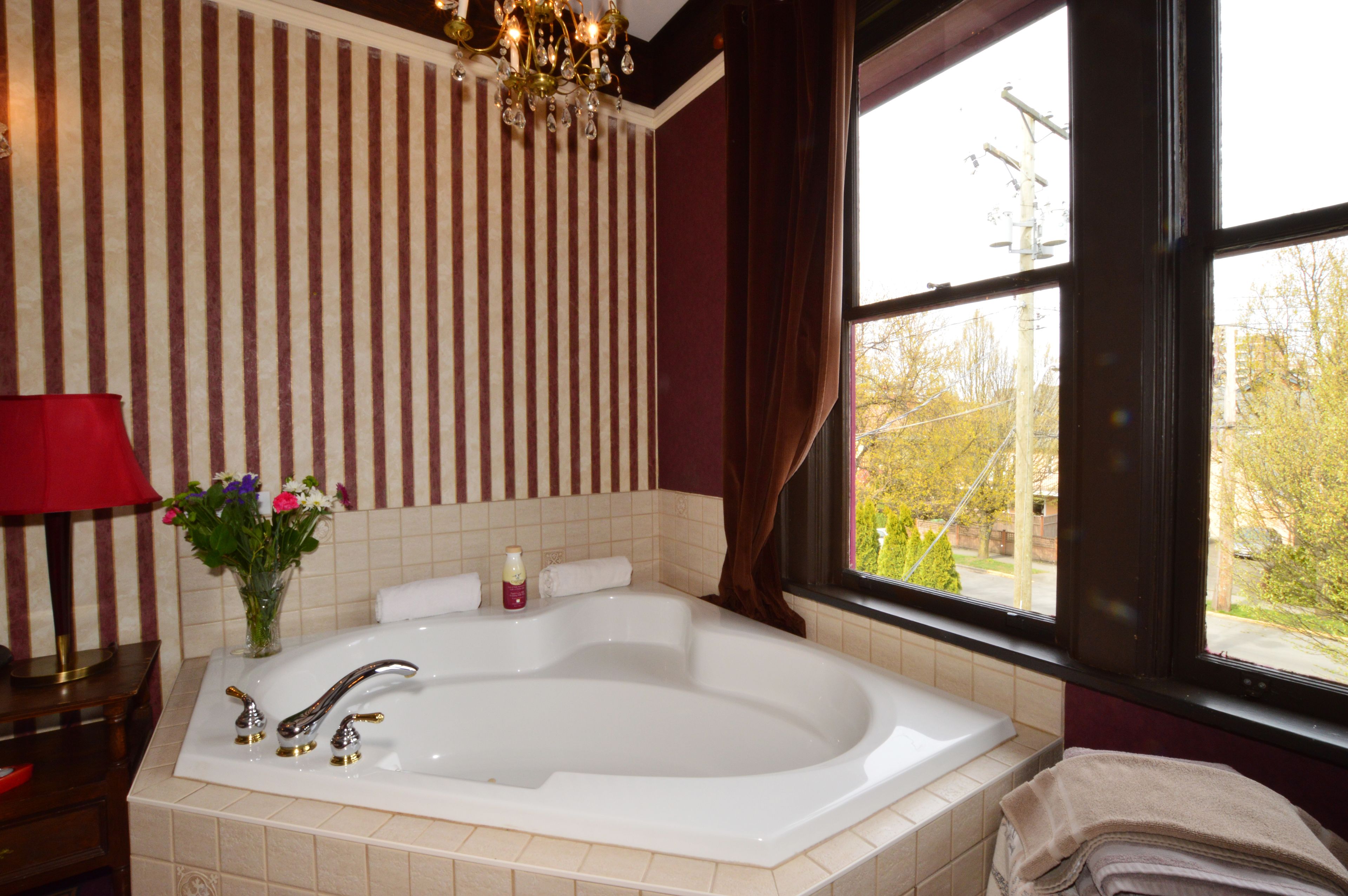 A bathroom with a large bathtub, a red lamp, a vase with flowers, and a window with a view of trees and a street.