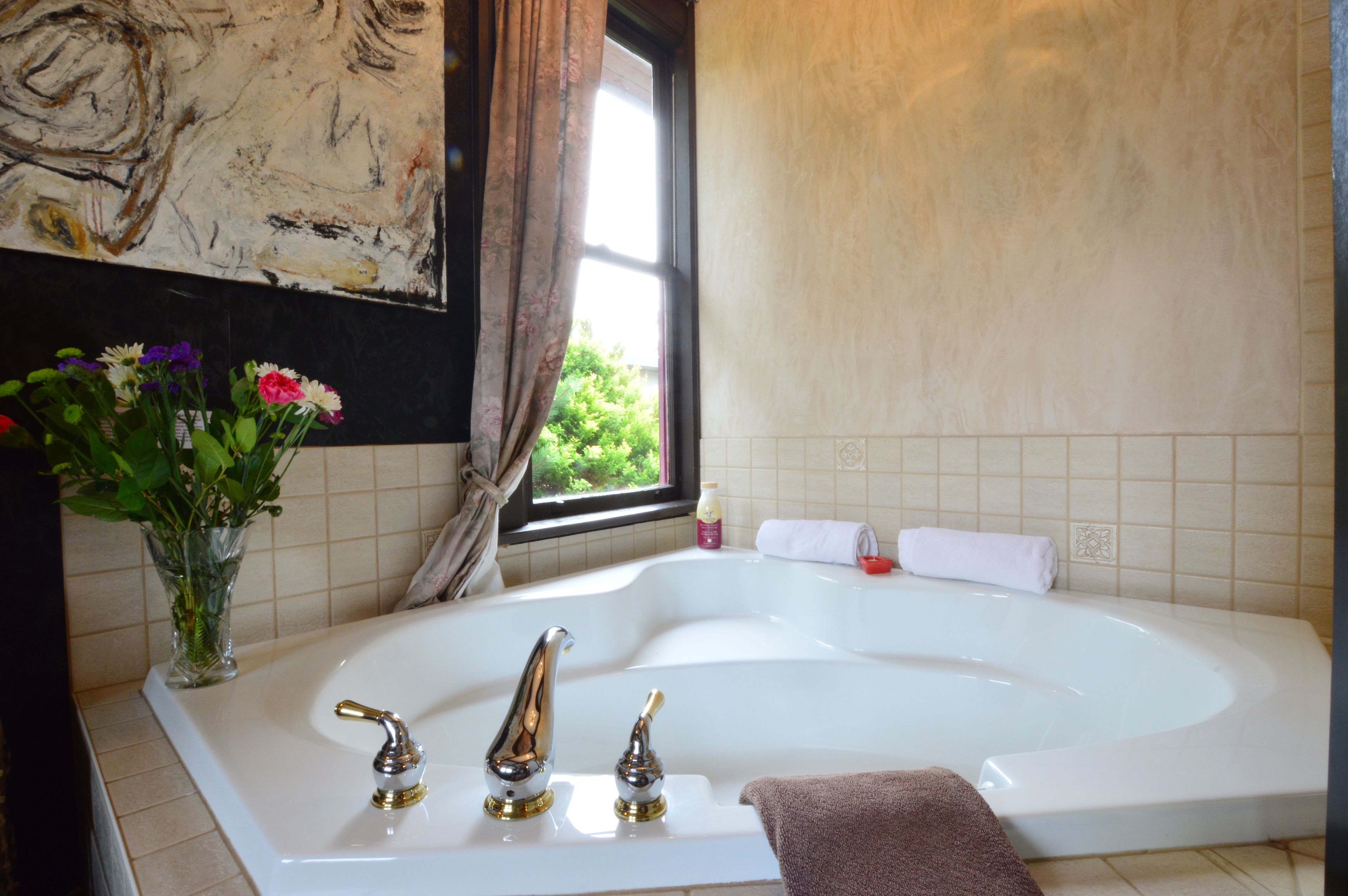 Ein Badezimmer mit einer großen Badewanne, einem Fenster mit Vorhängen, einer Vase mit Blumen und einem Handtuchhalter.