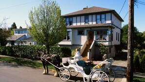 Un carruaje blanco tradicional con un asiento para pasajeros y un caballo atado, estacionado en una calle frente a una casa de dos pisos con un porche delantero y una escalera que lleva a la entrada.
