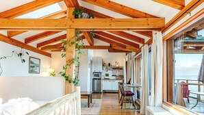 Interior de una cabaña con techo de madera, zona de comedor, cocina y un gran ventanal con vista al mar.