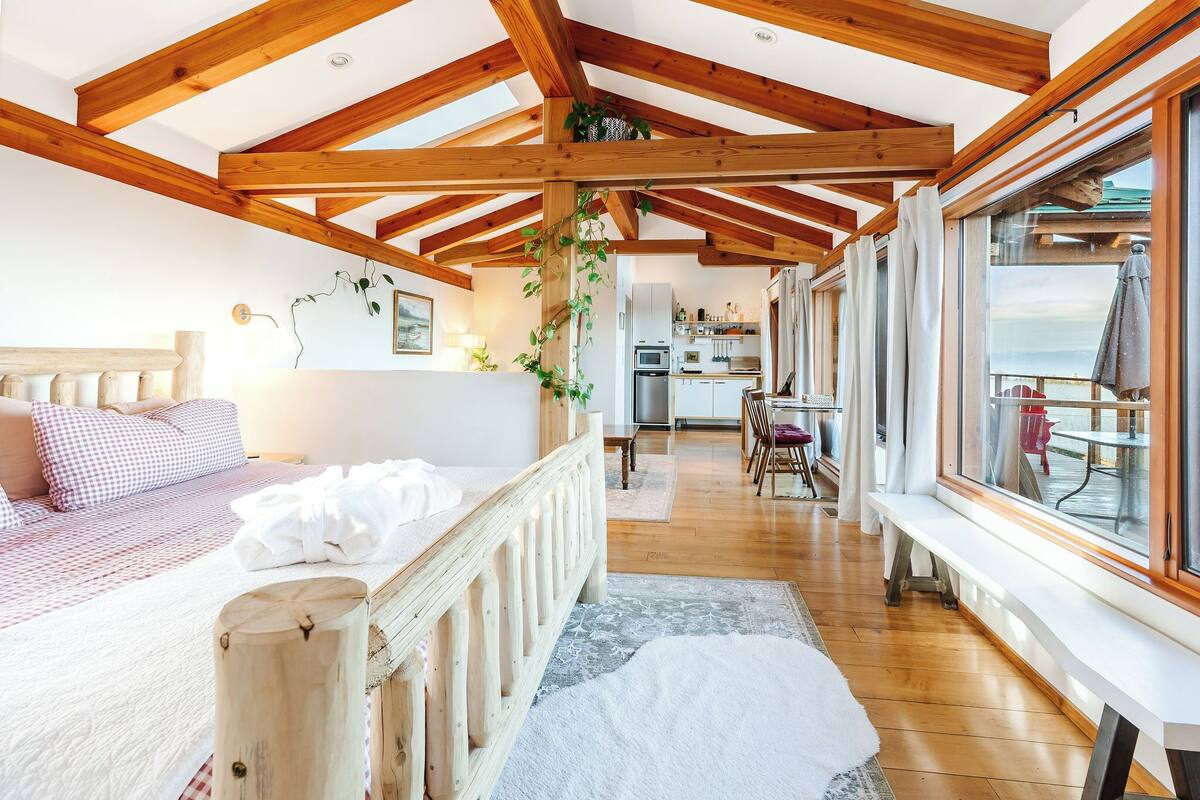 A bedroom with a wooden ceiling, a bed with a checkered bedspread, a bedside table, and a window with curtains.