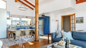 Amplia sala de estar con vigas de madera a la vista, un candelabro, un sofá y un comedor con una cocina al fondo.