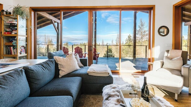 Una sala moderna con un sofá gris, un sillón blanco y una mesa de centro de vidrio. Se observa una estantería llena de libros y objetos decorativos. Grandes puertas de cristal dan a una terraza exterior con vistas a árboles y montañas.
