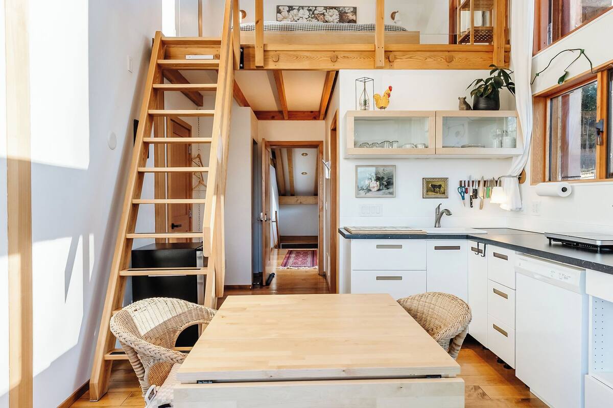 A compact, multi-level room with a wooden loft bed, a kitchen area, and a dining space.