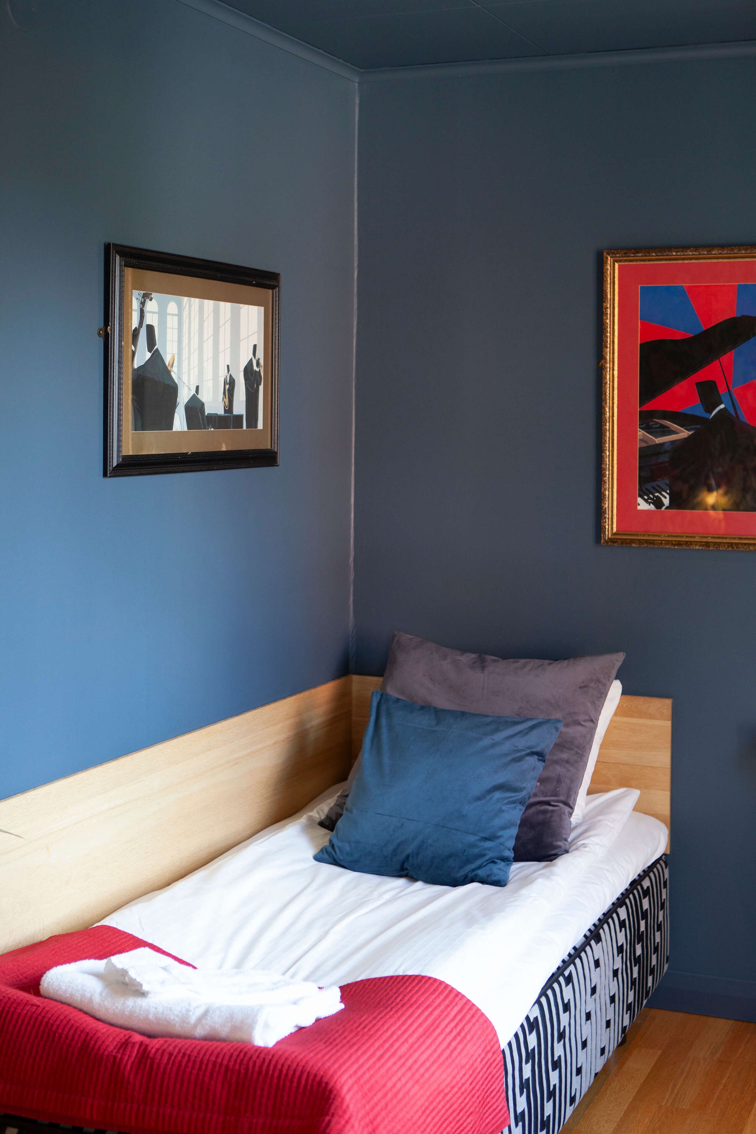 Una cama individual con cabecera de madera, ropa de cama blanca y dos almohadas. Hay dos cuadros en la pared, uno que muestra un grupo de personas y otro con un diseño abstracto en rojo y azul.