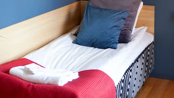 Uma cama de solteiro com cabeceira de madeira, roupa de cama branca e duas almofadas. Há duas imagens emolduradas na parede, uma retratando um grupo de pessoas e outra com um design abstrato em vermelho e azul.